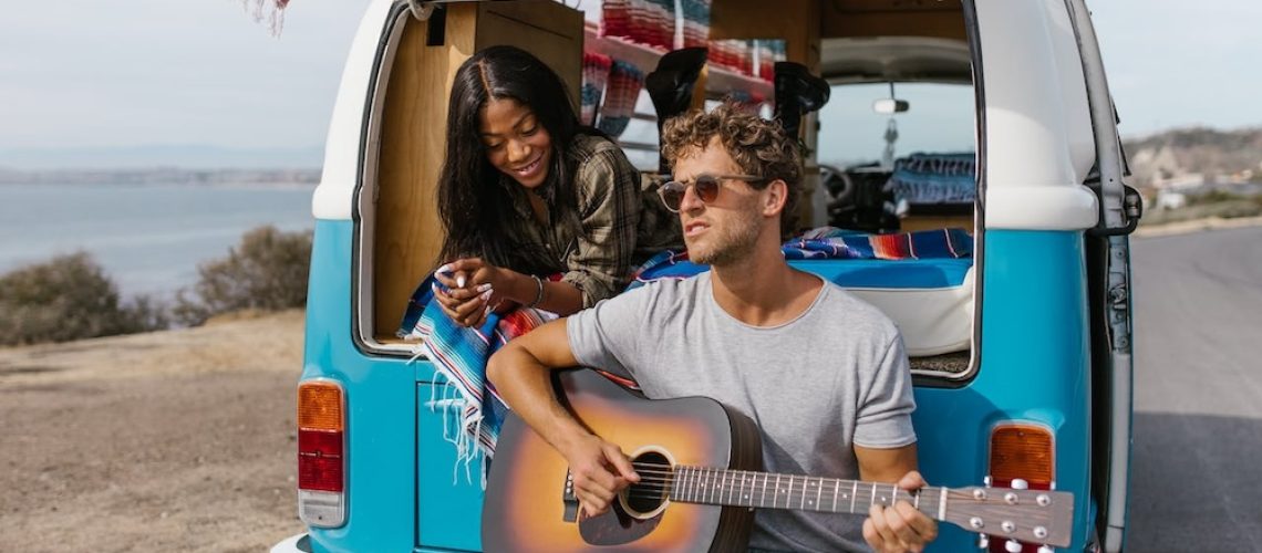 man playing the guitar while sitting on the bumper of a VW bus with a woman leaning over his shoulder listening